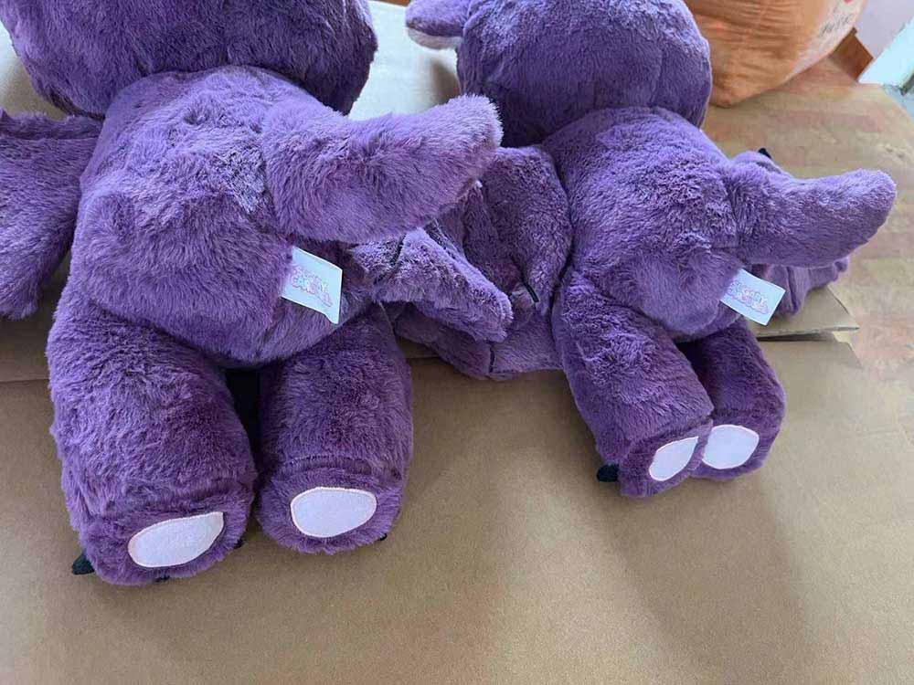 Several purple plush animal toys are shown from the back, lying on a flat surface. The toys feature soft, fluffy fabric, small pastel paw pads, and white care labels attached to their sides. The photo focuses on the stitching, texture, and placement of the labels on the plush toys.