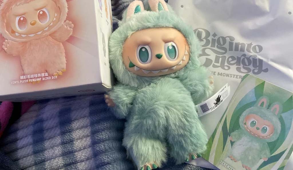 A mint-green Biginto Energy Labubu plush figure displayed with its original blind box, branded shopping bag, and character card. The Labubu features fluffy fur, bunny ears, large teal eyes, and a signature mischievous grin, shown resting on a soft blanket.