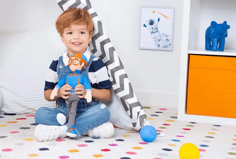A young boy sitting on a colorful polka-dot rug in a bright playroom, smiling while holding a plush doll dressed in blue and orange; playful décor and toys surround him, creating a cheerful and cozy scene.