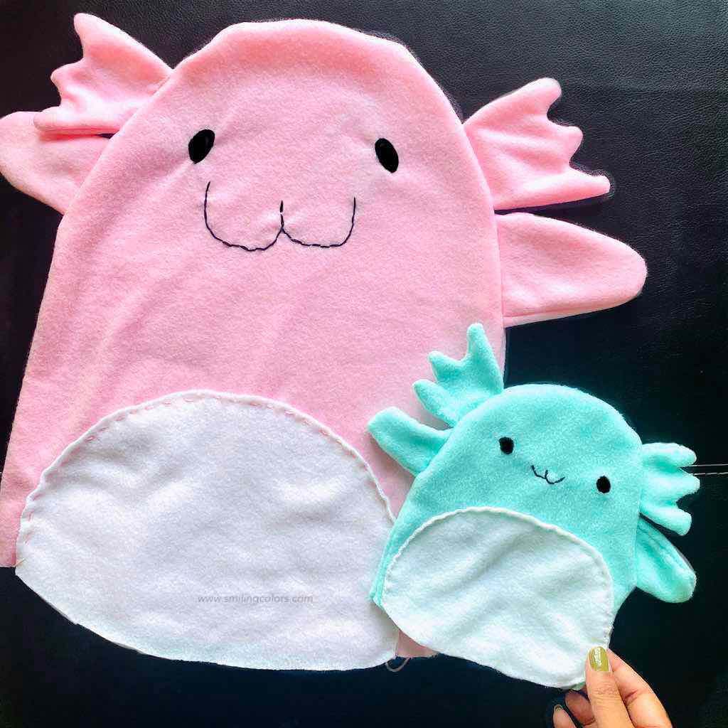 Handmade pink and teal axolotl plush toys made from soft fleece fabric, featuring stitched faces and white belly panels, displayed flat on a dark background.