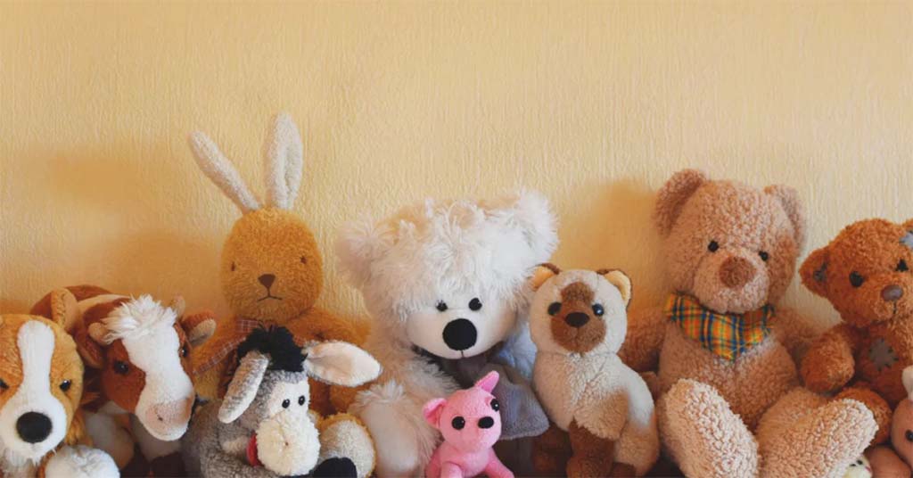 A row of various plush toys arranged against a yellow wall, including stuffed bears, a bunny, a cow, a donkey, a dog, and a small pink cat. The toys feature different textures and colors, creating a warm and cozy display.