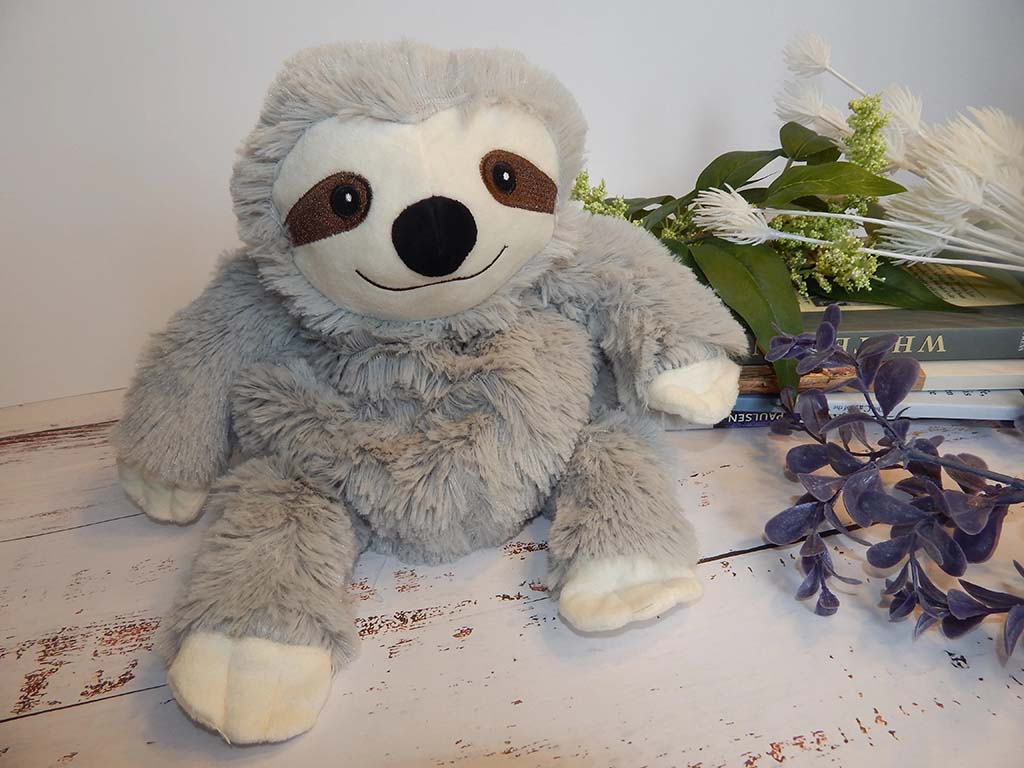 Cute grey sloth plush toy sitting on a rustic wooden surface with decorative greenery and books in the background. The sloth has a friendly embroidered face, long limbs, and fluffy faux-fur fabric, making it ideal for kids or collectors.