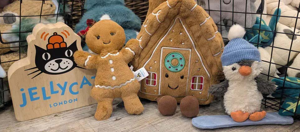 A festive display featuring Jellycat plush toys, including a gingerbread figure, a gingerbread house, and a penguin in a blue hat, arranged beside a wooden Jellycat sign.