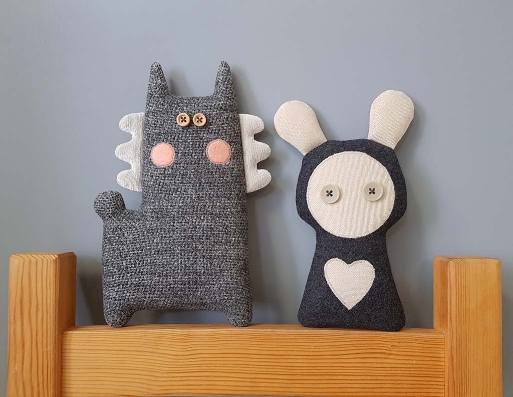 Two handmade minimalist plush toys sitting on a wooden bed frame, including a grey winged cat with button eyes and a dark plush bunny with long ears, button eyes, and a heart patch, against a light grey wall.