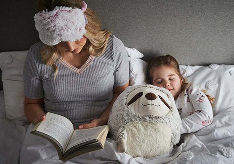 A mother reads a book in bed while her young daughter sleeps beside her hugging a soft sloth plush toy, creating a cozy bedtime scene.