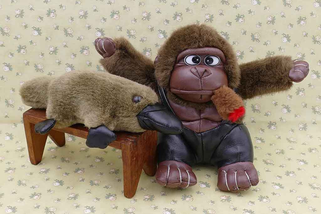 Vintage-style gorilla plush toy with faux leather face and limbs sitting beside a plush platypus on a small wooden bench against a floral patterned background.