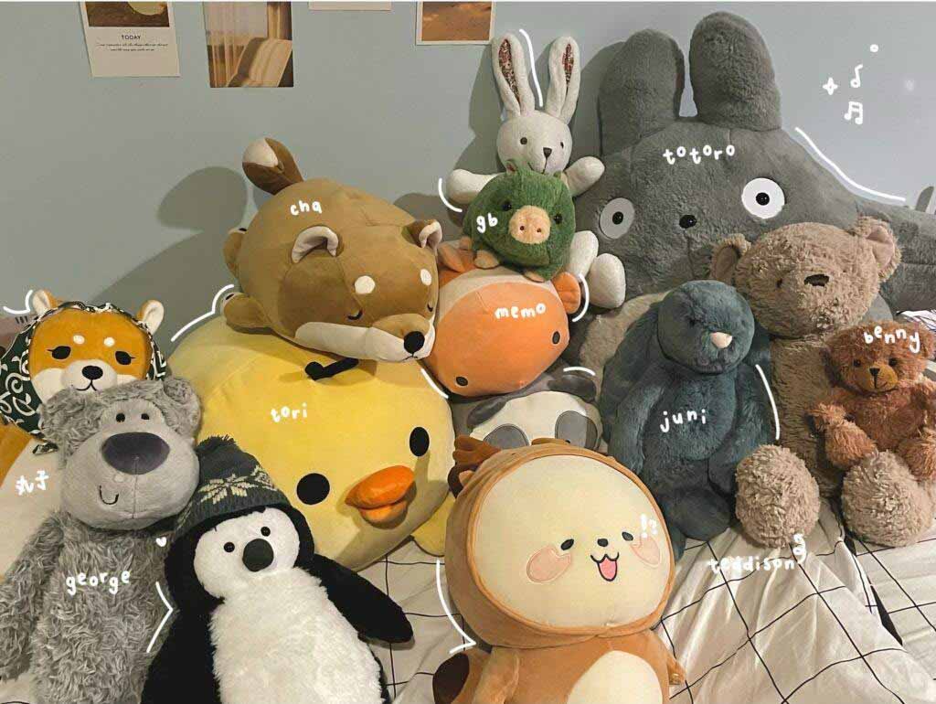 A cozy bedroom scene featuring a large collection of adorable plush toys, including bears, bunnies, penguins, foxes, and character plushies like Totoro, all arranged together on a bed with handwritten name labels for each stuffed animal.