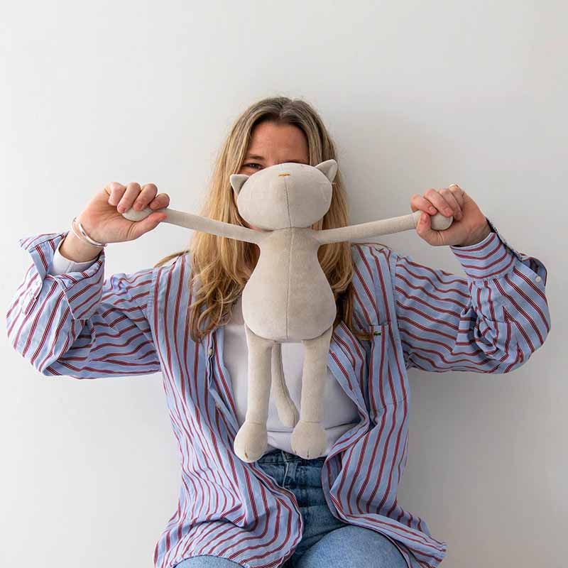 A person in a striped shirt holds a soft plush cat toy by its arms, raising it playfully in front of their face against a plain white background.