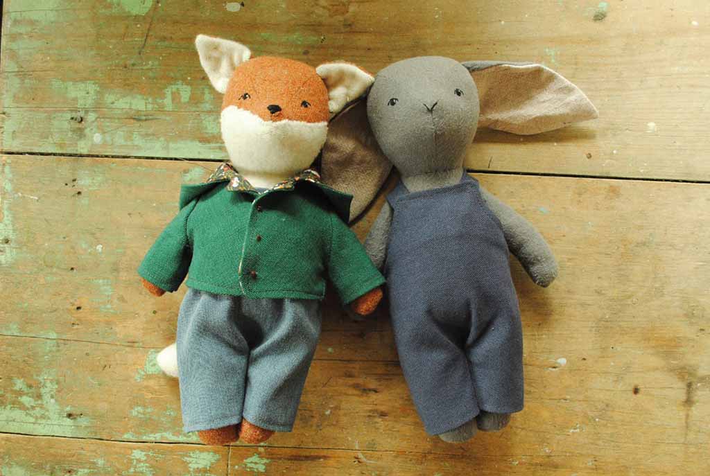 Two handmade cloth dolls—a fox in a green shirt and trousers and a bunny in blue overalls—lying side by side on a rustic wooden surface.