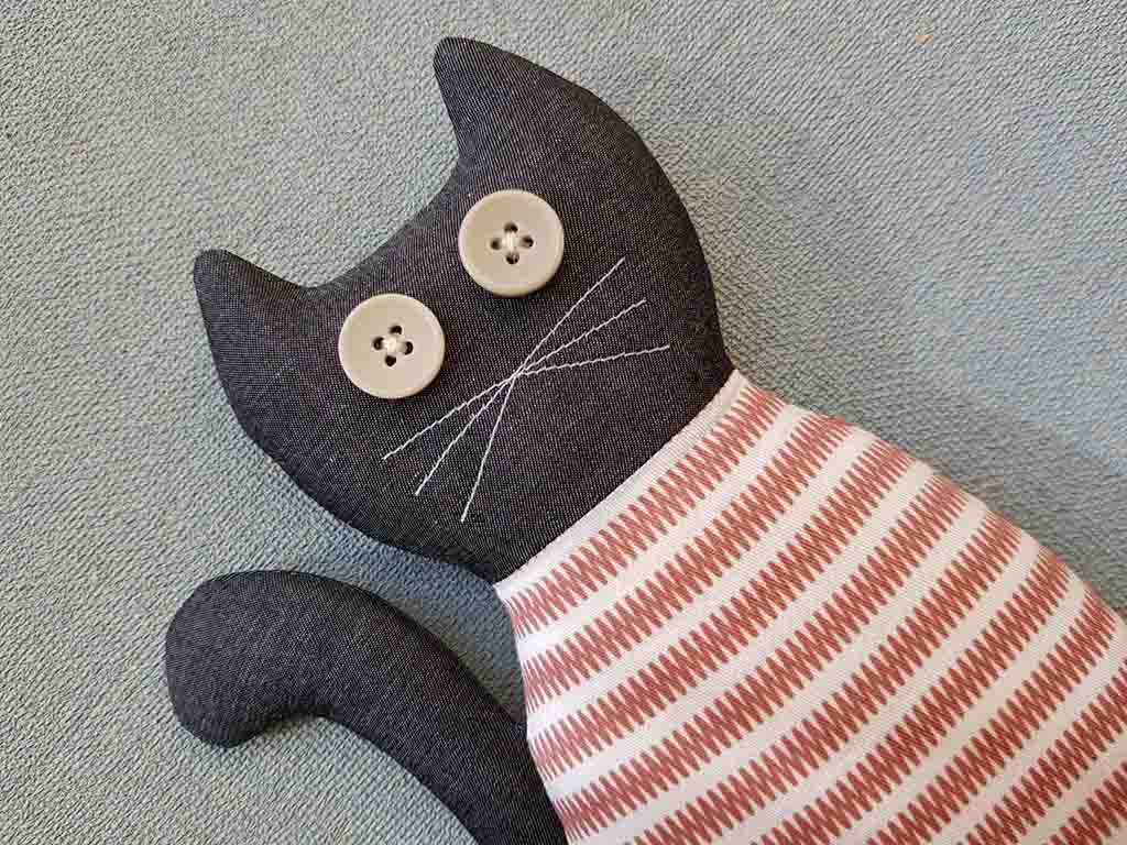 Handmade fabric cat plush with button eyes, embroidered whiskers, black head and tail, and a red-and-white striped body lying on a textured grey surface.