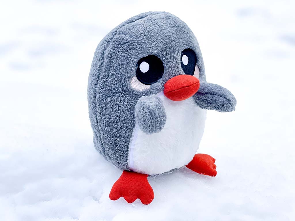 Adorable grey and white penguin plush toy with red beak and feet standing on fresh white snow, showing soft minky fabric and embroidered eyes.