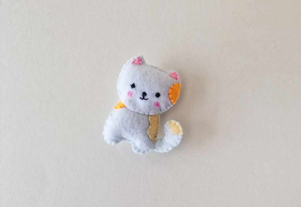 Cute handmade felt cat plush featuring grey body, embroidered face, pink cheeks, and yellow ear and tail details, displayed on a light background.
