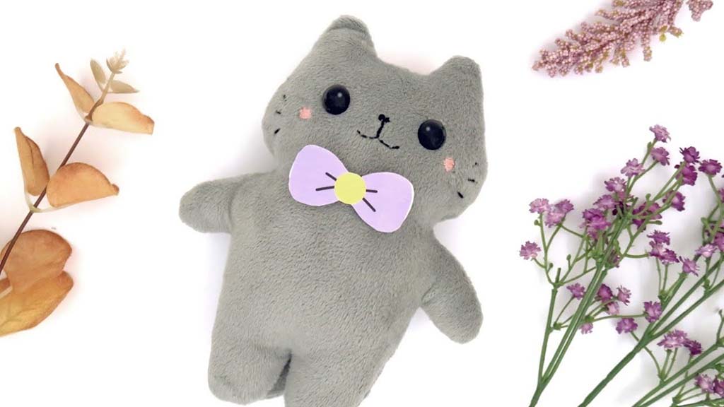 Soft grey cat plush toy with a lavender and yellow felt bow tie, embroidered face details, and rounded limbs, styled on a white background with dried leaves and purple flowers.