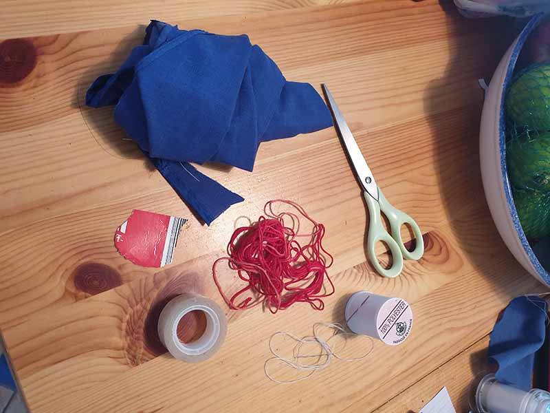 A collection of basic DIY sewing materials on a wooden table, including blue fabric scraps, scissors, thread, red elastic bands, tape, and small craft supplies arranged for a sewing project.