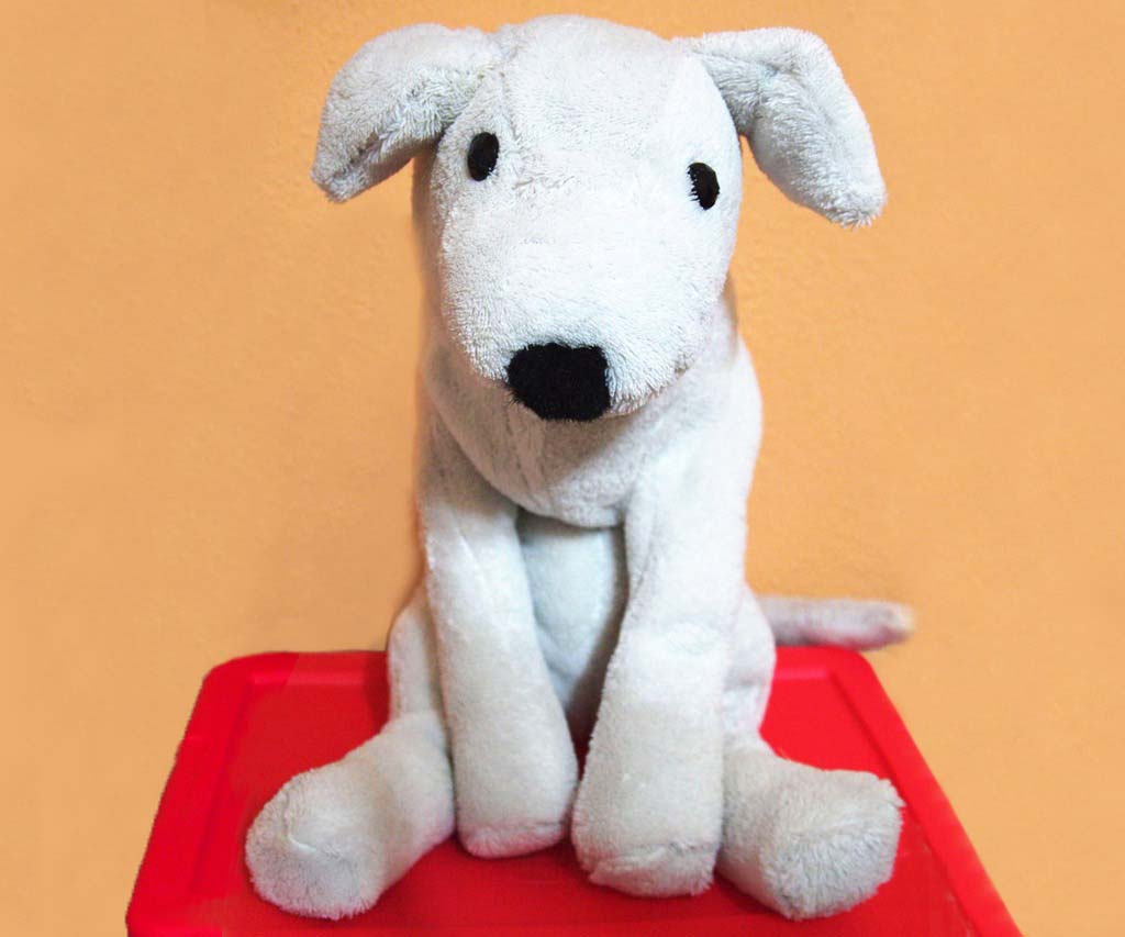 White plush dog toy sitting on a red surface against a warm orange background, with floppy ears and black stitched facial features.
