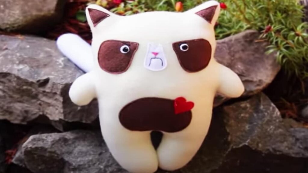 Handmade grumpy cat plush toy with brown felt patches and a small red heart, displayed outdoors on rocks.
