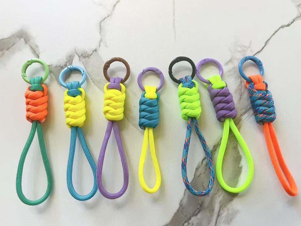 Colorful handmade paracord loop keychains arranged in a row, featuring braided knots and matching plastic rings in bright neon colors, displayed on a marble background.