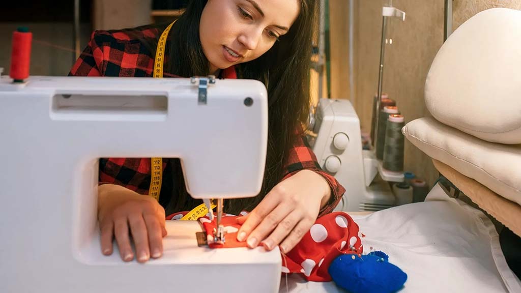 Woman using a sewing machine to stitch colorful plush toy fabric, with measuring tape around her neck and sewing tools nearby.