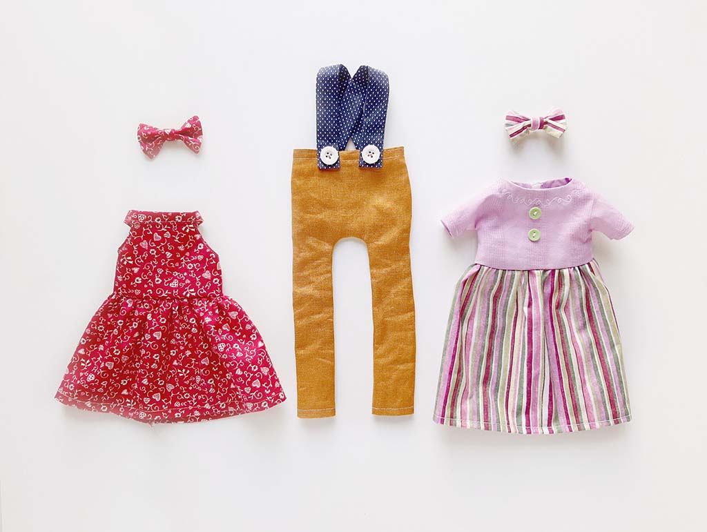 Assorted handmade doll or plush toy clothes, including dresses, overalls, bow ties, and striped outfits, neatly arranged on a white background.