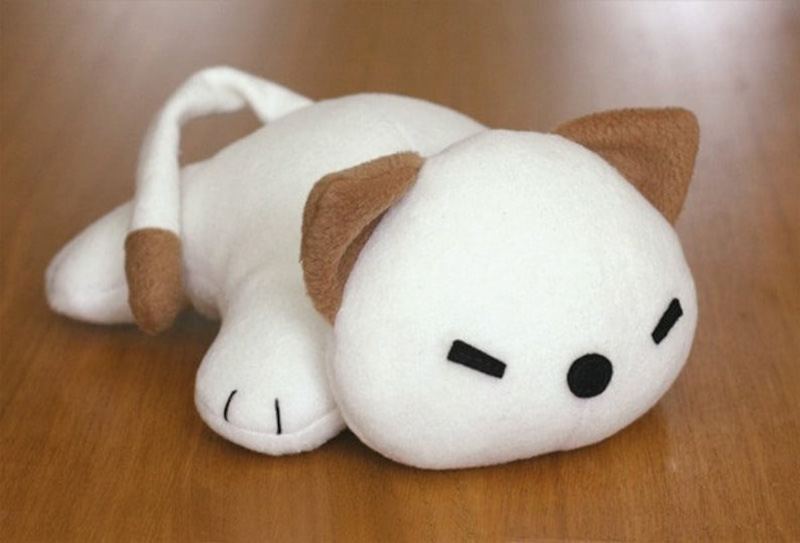 Simple white sleeping cat plush with brown ears, tail tip, and paw details, lying on a wooden surface in a minimalist kawaii style.