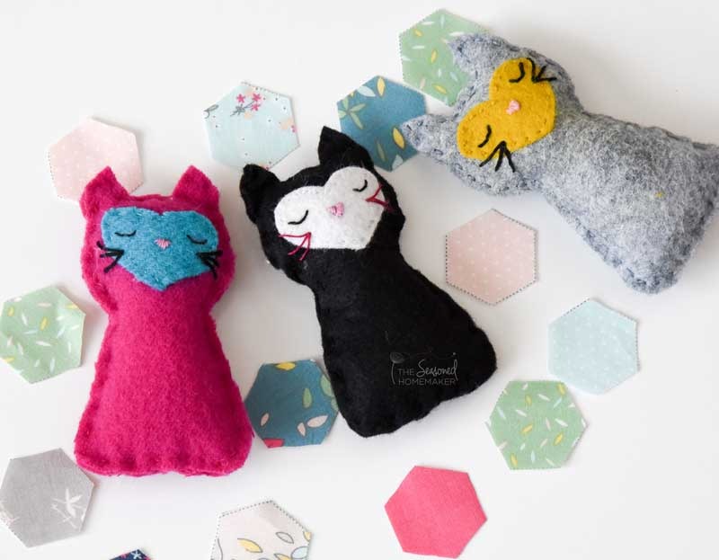 Three handmade felt cat plushies in pink, black, and gray colors, each with embroidered sleeping faces, surrounded by pastel hexagon fabric pieces on a white background.