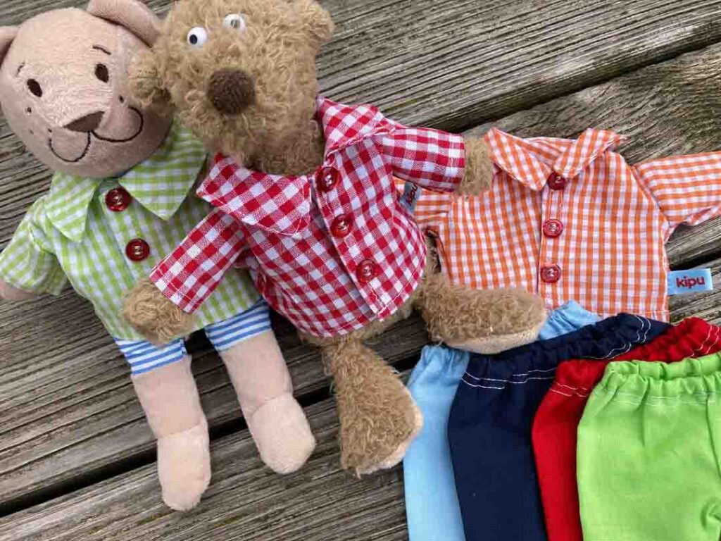 Two plush toy animals wearing green and red gingham shirts with buttons, displayed alongside additional doll-sized gingham tops and colorful elastic-waist pants on a wooden surface.