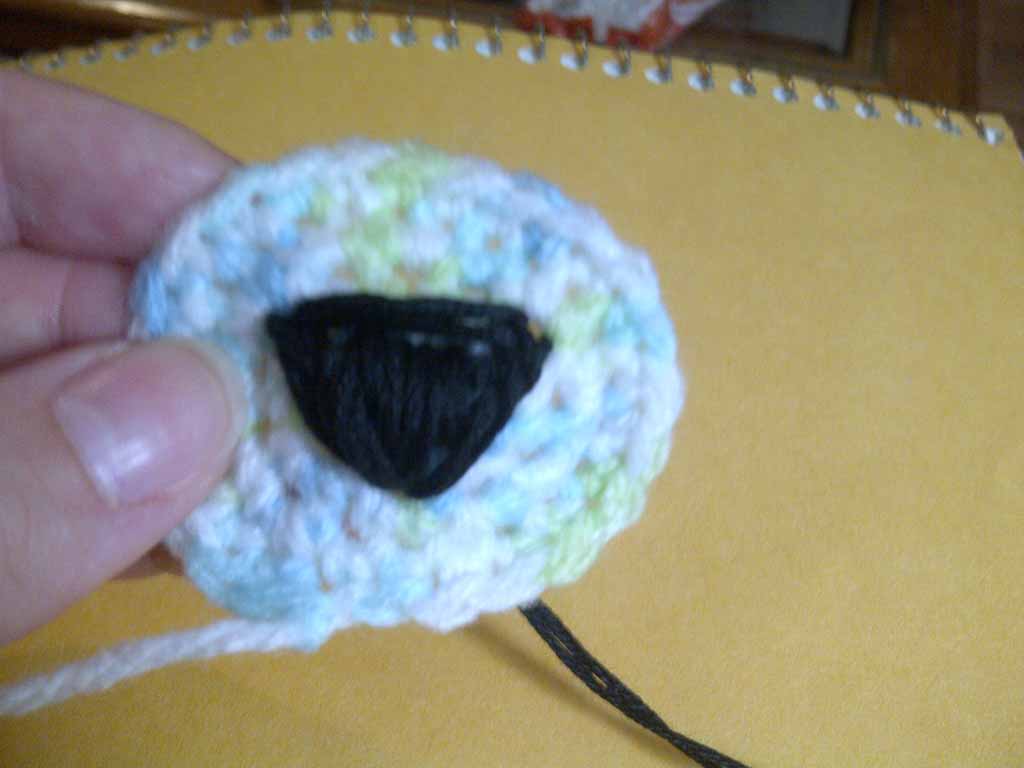 Close-up of a crocheted oval piece with pastel yarn and a black embroidered triangular eye patch, held in someone’s hand during plush toy making.