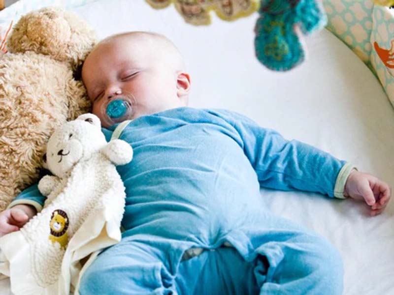 A baby in blue pajamas sleeps peacefully in a crib while holding a soft lovey blanket and stuffed bear, with a pacifier in their mouth.