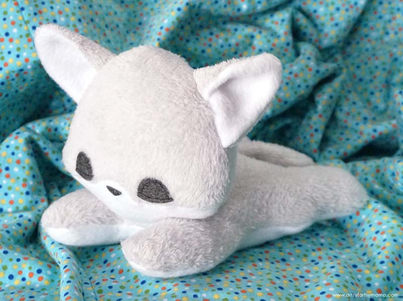 Soft grey cat plush toy with large ears and minimalist embroidered face, lying on a blue polka-dot fabric background.