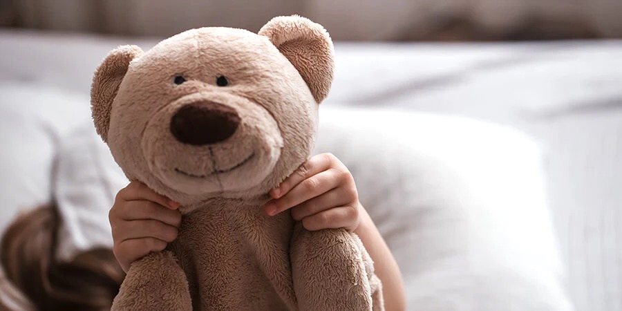 A child holding a soft beige teddy bear close to their face while sitting on a bed, showing a cozy and comforting moment.