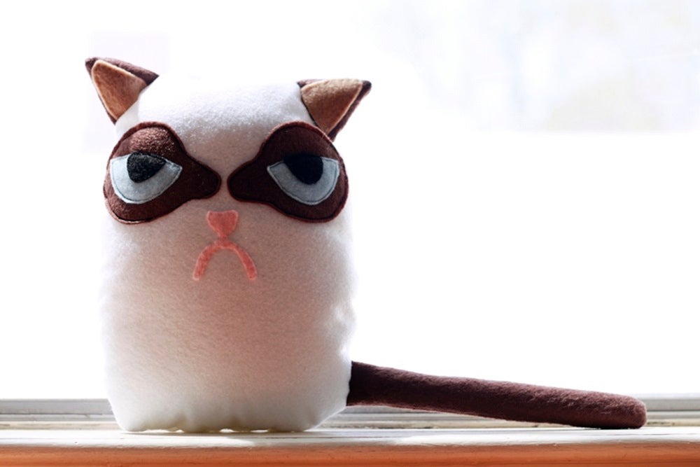 Cute grumpy-faced cat plush toy with brown ears, dark eye patches, and a long brown tail, sitting on a windowsill against a bright white background.
