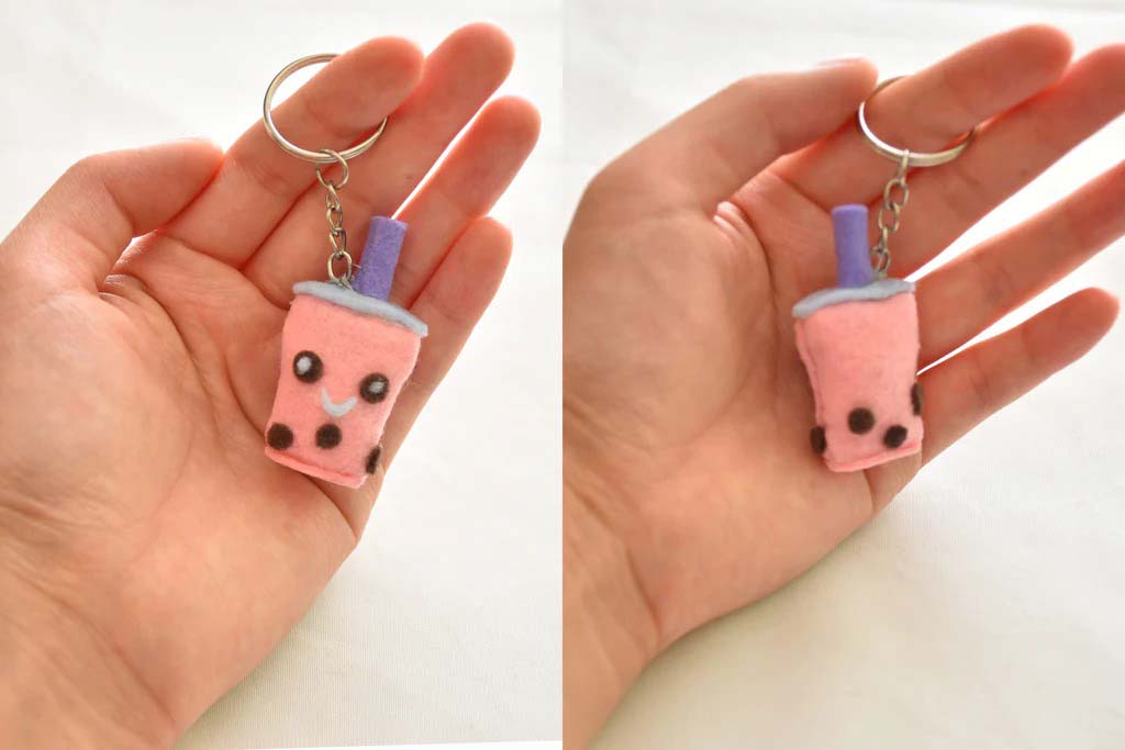 Hand holding a handmade pink felt boba tea keychain plush with a purple straw, embroidered smiley face, and brown tapioca pearls; front and back views shown.