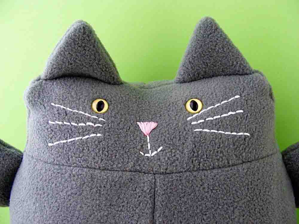 Close-up of a grey handmade cat plush with embroidered whiskers, pink stitched nose, and yellow button-style eyes against a green background.