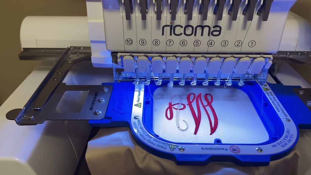 Ricoma multi-needle embroidery machine stitching a red monogram design inside a blue hoop, showing precise thread control and professional embroidery setup.