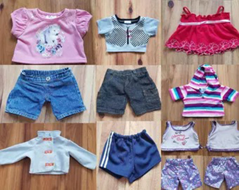 A collection of assorted doll and stuffed animal clothes laid out on a wooden floor, including shirts, shorts, hoodies, dresses, and patterned outfits in various colors and styles.