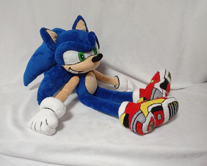 Soft plush toy of Sonic the Hedgehog sitting on a white surface, featuring blue fur, green eyes, white gloves, and oversized red, yellow, and black shoes.