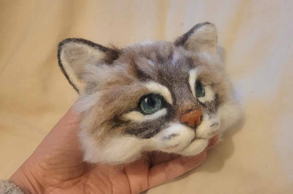 Hand holding a realistic needle-felted cat head made of wool, showing detailed fur texture, blue eyes, and soft sculpted features.