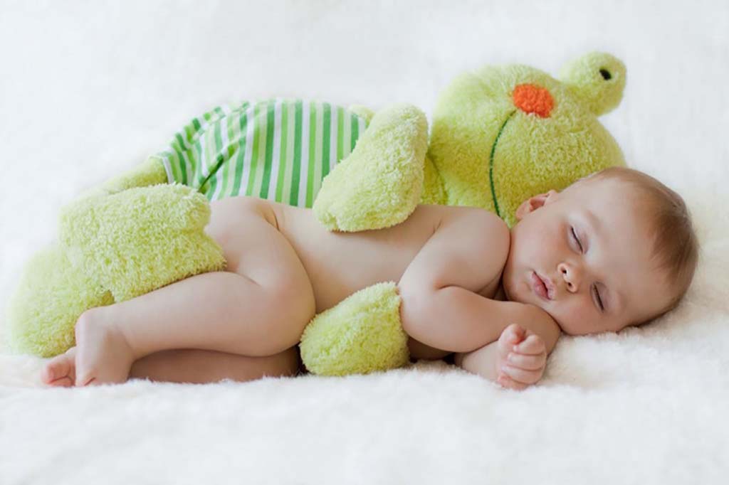 A sleeping baby lies on a soft white surface while hugging a large green plush toy, resting peacefully with eyes closed.