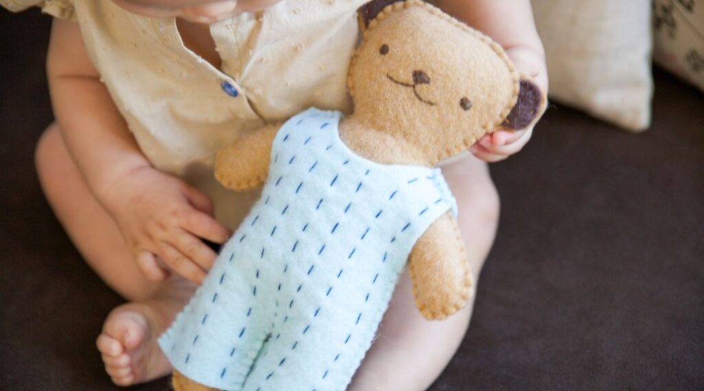 A baby sitting on the floor gently holding a handmade felt teddy bear wearing a light blue stitched outfit.