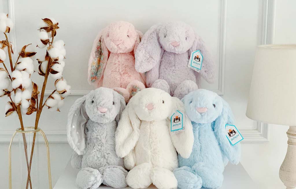 Group of pastel-colored Jellycat bunny plush toys arranged on a white shelf, featuring soft long ears and embroidered details.