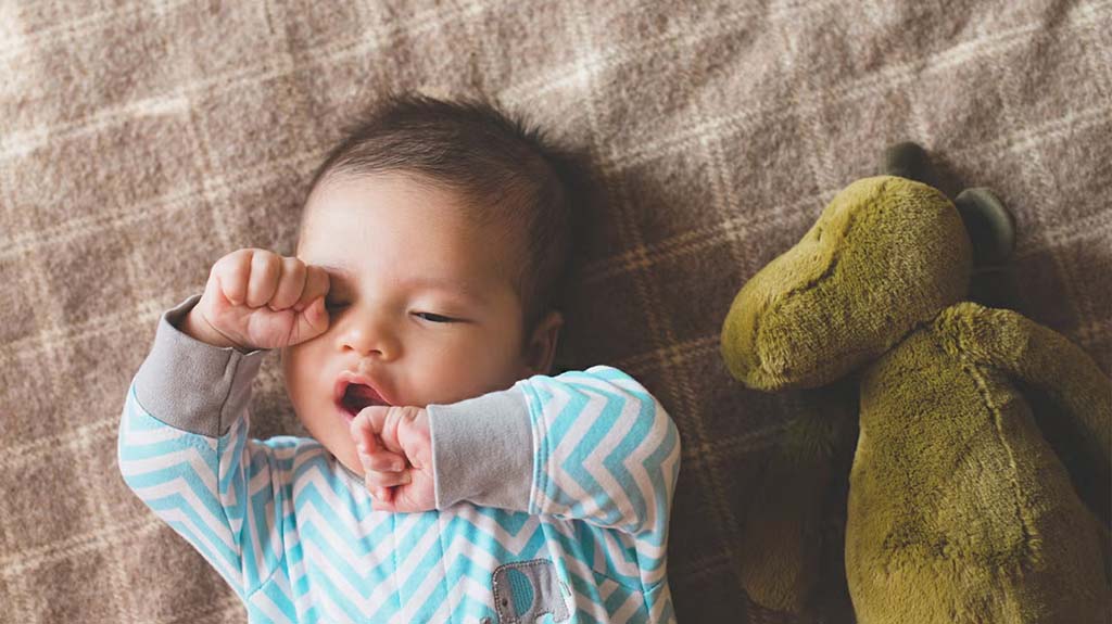 A baby in a blue patterned onesie yawns while lying on a blanket next to a green stuffed animal.