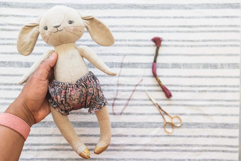 A person holding a handmade fabric bunny doll wearing floral shorts, placed on a striped surface beside embroidery floss and small sewing scissors, illustrating a plush-making or hand-sewing process.