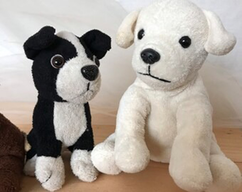 Two plush dog toys—one black-and-white with button eyes and one solid white with stitched facial features—sitting side by side on a wooden surface with a soft background.