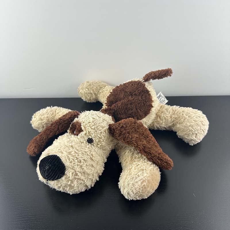 A floppy plush dog toy with a cream body, brown ears, brown patches, and a soft textured fabric, lying flat on a black surface against a light gray background.