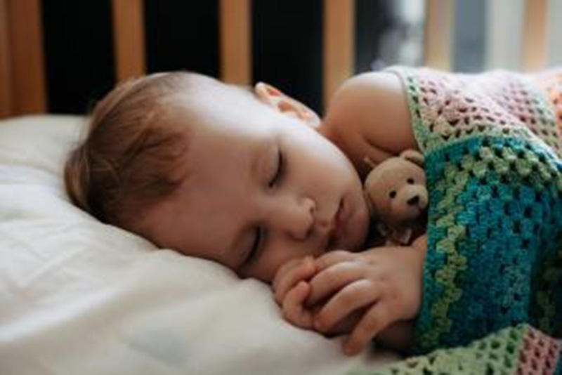 A baby sleeps peacefully in a crib, wrapped in a colorful crochet blanket while holding a small teddy bear close to their chest.