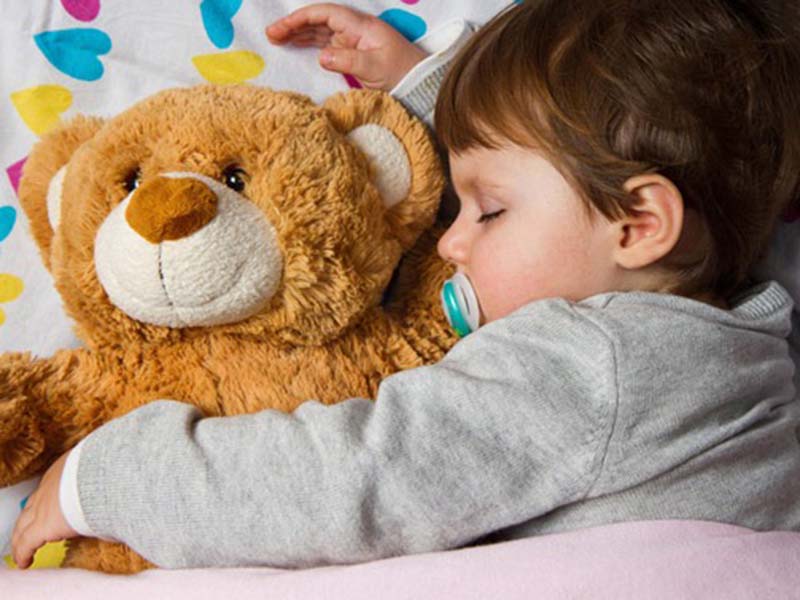 A toddler sleeps peacefully while hugging a soft brown teddy bear, holding a pacifier in their mouth and resting on a colorful heart-patterned blanket.