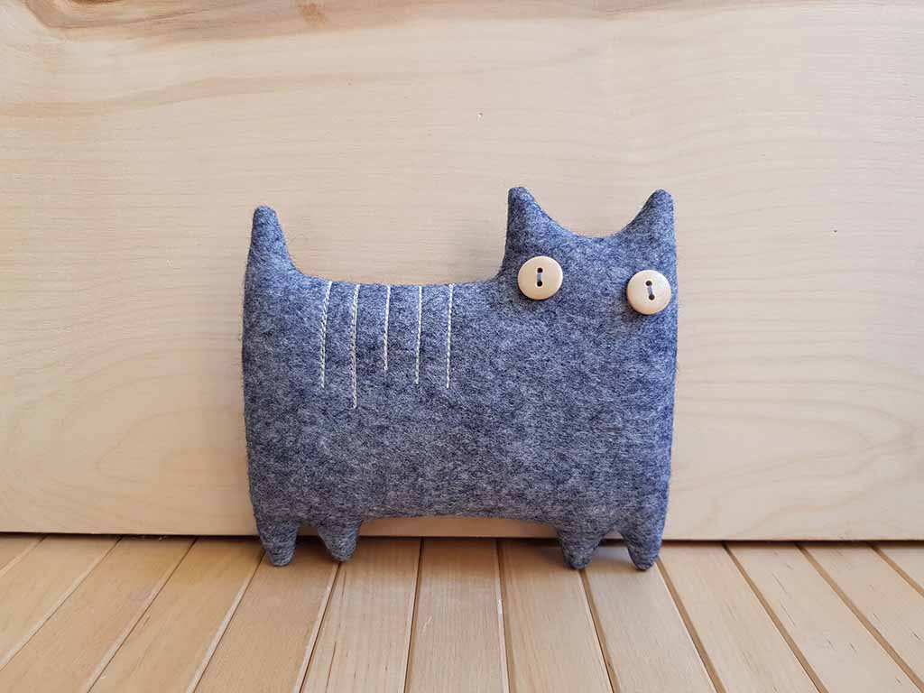 Handmade minimalist grey felt cat plush with button eyes and simple embroidered details, standing against a wooden background.