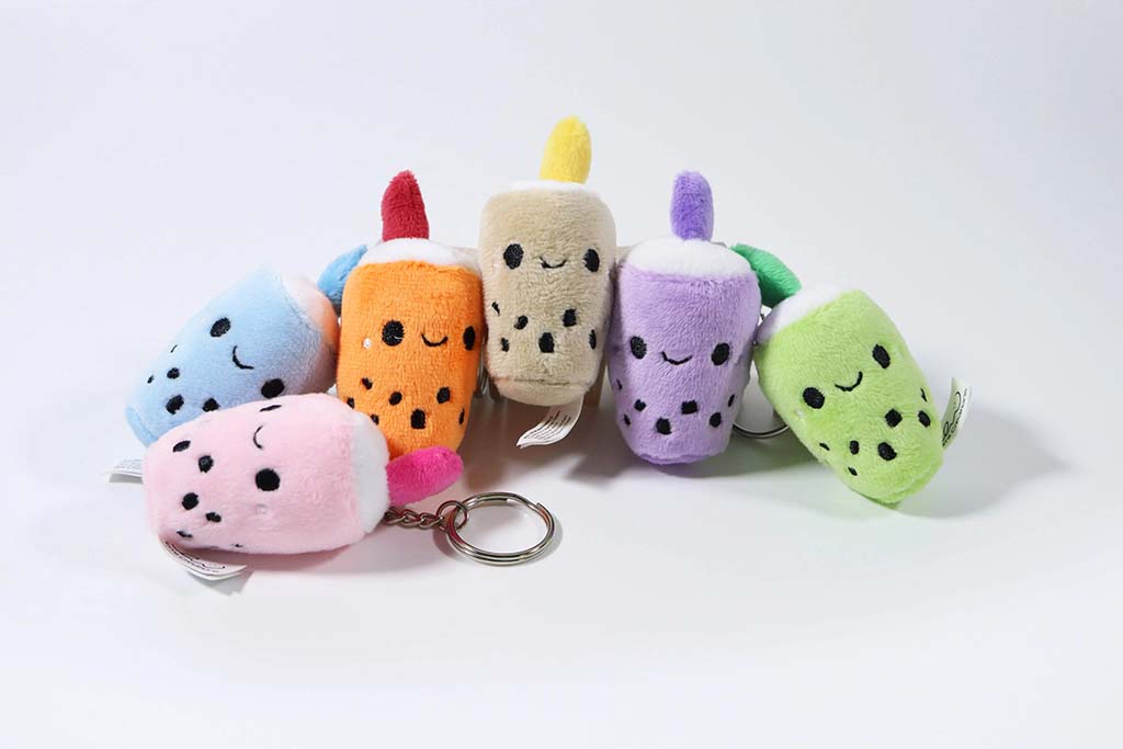 Six colorful boba tea plush keychains arranged in a row, each with smiling embroidered faces, bubble details, and soft pastel colors including pink, blue, orange, beige, purple, and green.