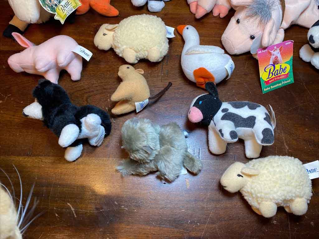 A collection of small farm-themed plush toys, including cows, sheep, pigs, dogs, geese, and other animals, arranged on a wooden surface with visible product tags.