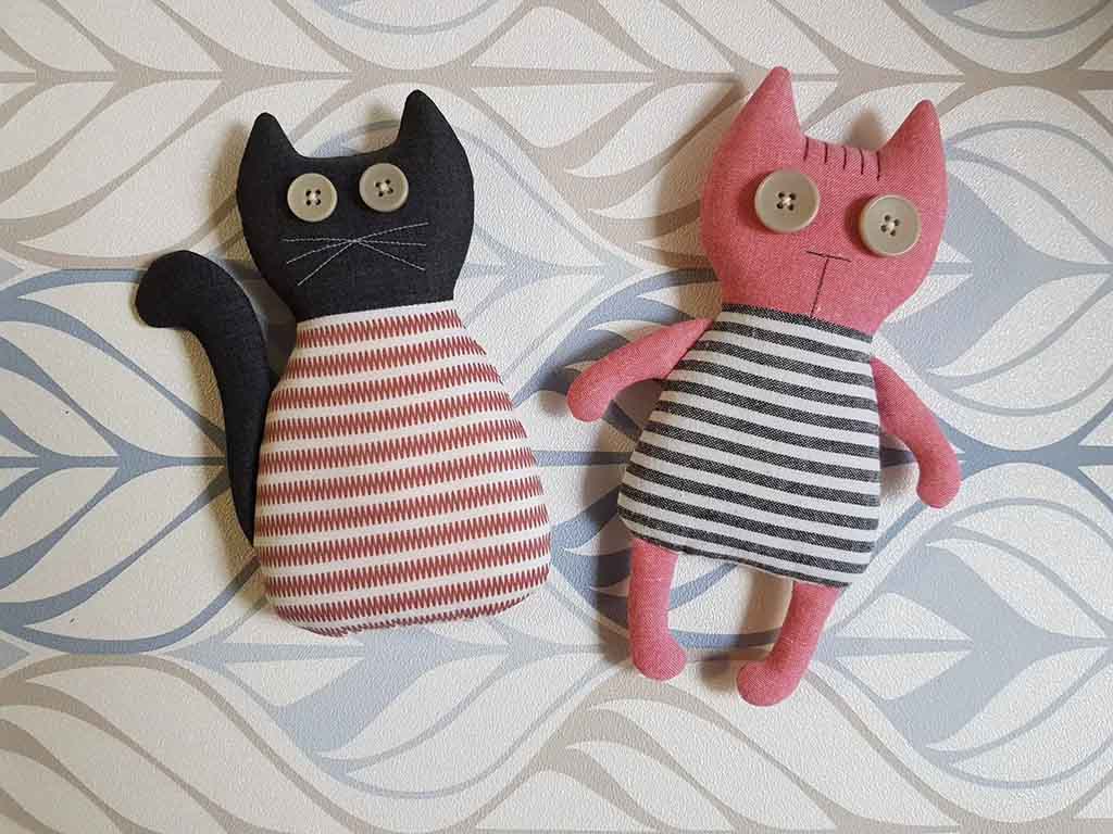 Two handmade fabric cat plushies with button eyes, one black with a red patterned body and one pink with a striped dress, displayed on a decorative background.